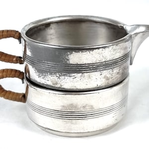 May include: A silver creamer with a double-walled design and a woven brown handle. The creamer has a smooth, polished finish and a slightly worn appearance.