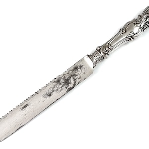 Puede incluir: Un cuchillo para pastel de plata con un mango adornado y una hoja dentada.