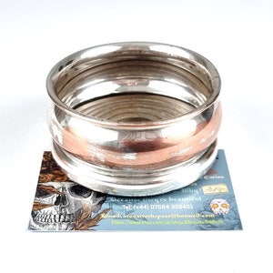 May include: A silver napkin ring with a copper band around the middle. The ring is made of multiple layers of metal and has a shiny finish.