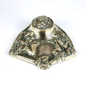 May include: A silver-colored inkwell with a triangular base and a round top. The inkwell is decorated with detailed leaf and berry patterns. The top has a textured design, and the base has a small, decorative foot.