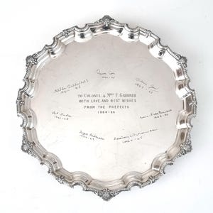 May include: A silver-colored, ornate serving tray with a scalloped edge. The tray has engraved text and signatures, including "TO COLONEL & MRS F GARDINER WITH LOVE AND BEST WISHES FROM THE PREFECTS 1964-65". The tray is on a white background.