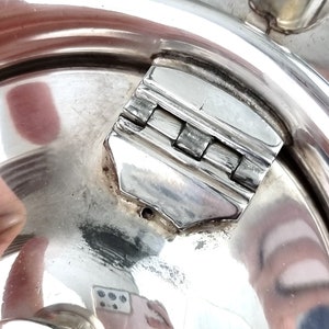 May include: Close-up of a silver teapot lid with a hinged latch. The latch is made of metal and has a polished finish.