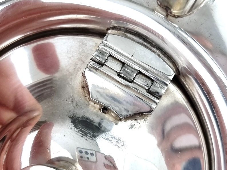 May include: Close-up of a silver teapot lid with a hinged latch. The latch is made of metal and has a polished finish.