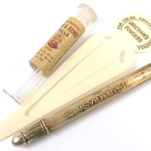 May include: A vintage silver-colored metal pen with the inscription "SANITAS" on the side. The pen is lying on a white plastic surface with a glass vial and a white paper tag with the text "FOR COLDS. CHILLS AND INFLUENZA BEECHAM'S POWDERS" on it.
