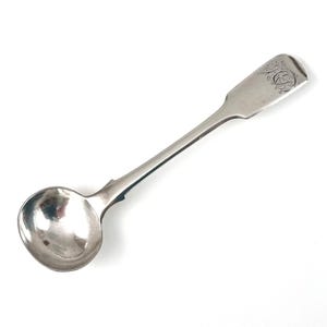 Könnte beinhalten: Ein silberner Löffel mit einer runden Schüssel und einem langen, schlanken Stiel. Der Griff hat ein rechteckiges Ende mit einem eingravierten Design. Der Löffel ist wahrscheinlich antik oder vintage und vor einem weißen Hintergrund platziert.