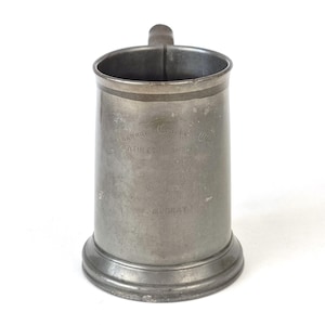 Könnte beinhalten: Ein Vintage-Zinnkrug mit Henkel. Der zylindrische Körper weist eingravierten Text mit der Aufschrift "ATHLETIC SPORTS" und "W. MURRAY" auf. Der Krug hat einen leicht ausgestellten Boden und ein Band um den oberen Rand. Die Gesamtfarbe ist ein gedämpftes Grau.