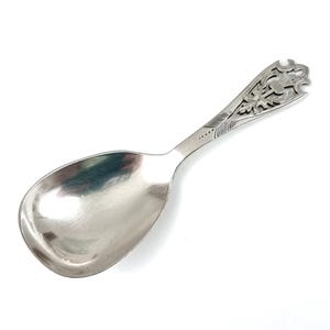 May include: A silver-toned metal spoon featuring a wide, rounded bowl and a decorative handle. The handle displays an intricate cutout design depicting a thistle motif, with fine engraved lines extending from the bowl toward the patterned top.