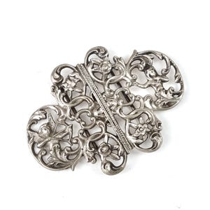 Puede incluir: Una hebilla de cinturón de metal plateado con un intrincado diseño calado. El patrón de filigrana incluye motivos florales y una figura de querubín sosteniendo un arco y una flecha. La barra vertical central tiene un acabado texturizado.