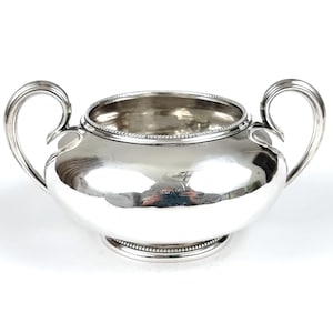 May include: A shiny silver sugar bowl with two curved handles. The bowl has a rounded body and a beaded rim and base. The interior is reflective, and the overall design is classic and elegant. The bowl is likely made of sterling silver.
