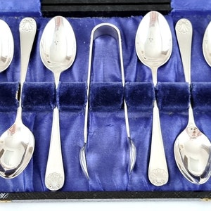 Puede incluir: Un juego de seis cucharaditas de plata y un par de tenazas para azúcar en una caja de madera forrada de terciopelo azul.