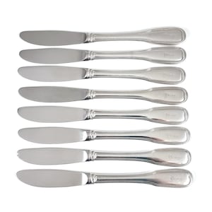 Può includere: Un set di otto coltelli da tavola in metallo color argento disposti in file parallele. Ogni coltello presenta una lama liscia e arrotondata e un manico con un sottile dettaglio a rilievo lungo i bordi, che termina con una punta arrotondata.