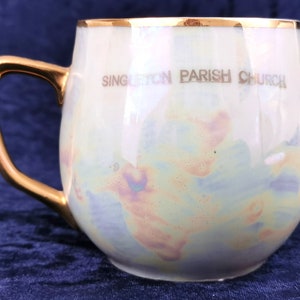 May include: A white ceramic mug with a gold rim and handle. The mug has a light blue, pink, and yellow marbled design and says "Singleton Parish Church" in gold lettering.