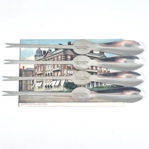 以下が含まれることがあります： 「クイーンズホテルサウシー」と刻まれた銀製のカクテルピックが5本。ピックは、クイーンズホテルサウシーのヴィンテージのポストカードの上に置かれています。