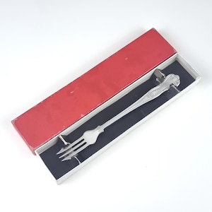 Op de afbeelding: Een zilveren serveervork met een sierlijk handvat, gepresenteerd in een rood-witte doos met een zwart interieur. De vork heeft drie tanden en een decoratief bloemmotief op het handvat.