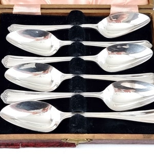 May include: Set of six silver teaspoons in a wooden box with a red velvet lining. The spoons are arranged in a row, with the handles facing up. The spoons are polished and shiny.