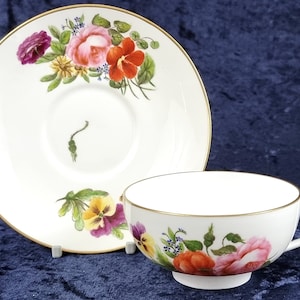 May include: A white teacup and saucer set with a gold trim. The saucer features a floral design with pink, red, yellow, and purple flowers. The teacup has a similar floral design with a pink, red, and yellow rose.
