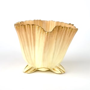 May include: A decorative, fan-shaped ceramic vase with a scalloped rim and three feet. The vase features a cream-colored base that transitions to a light peach hue towards the top. The rim and feet are accented with gold.