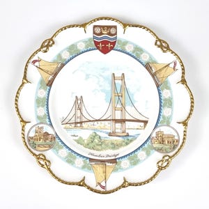 Puede incluir: Un plato decorativo blanco con un borde dorado, que presenta el puente Humber como imagen central. El plato incluye veleros, edificios y un borde floral. El texto "Humber Bridge" está debajo de la imagen del puente.