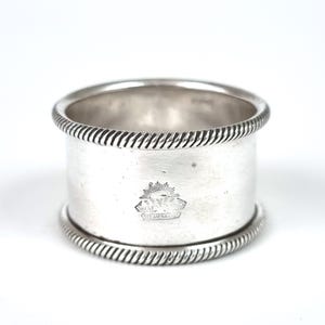 May include: A silver napkin ring with a rope-like design on the top and bottom edges. The ring has a cylindrical shape and a smooth, polished surface. A small emblem is visible on the front of the ring.