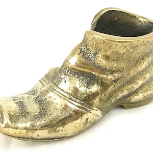 May include: A brass shoe-shaped trinket or decorative object. The shoe is detailed with a textured surface and a folded-over top.