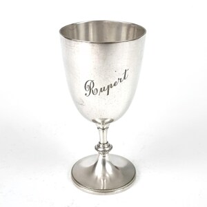 Peut inclure: Un gobelet en argent avec un large bol et une base à pied. Le mot "Rupert" est gravé avec élégance sur le bol. Le gobelet a une finition polie et un bord perlé décoratif sur la base. Une pièce classique pour servir des boissons.