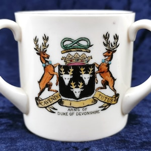 Puede incluir: Taza de porcelana blanca con dos asas, con un escudo de armas con dos ciervos, una corona y el texto "Cavendo Tutus" y "Arms of Duke of Devonshire".