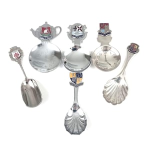 Könnte beinhalten: Eine Sammlung von fünf silberfarbenen Souvenir-Löffeln aus Metall. Zwei Löffel haben Zifferblätter in den Laffen, während zwei andere muschelförmige Laffen aufweisen. Jeder Griff ist mit einem farbigen Emaille-Wappen versehen, das Orte wie Firenze, Isle of Wight, Felixstowe und Bournemouth darstellt.