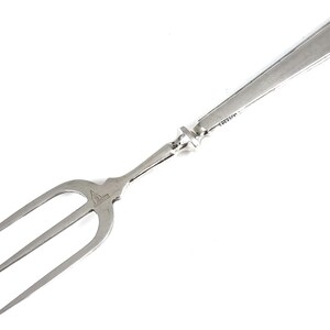 Op de afbeelding: Een zilveren serveervork met drie tanden. De handgreep is rechthoekig en loopt taps toe naar het uiteinde. De vork heeft een decoratief element waar de tanden de handgreep ontmoeten. Het woord "STERLING" is op de handgreep gestempeld.