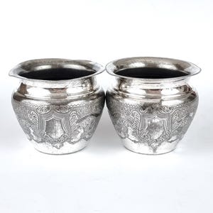 May include: Two silver cups with intricate floral designs etched into the surface. The cups are round and have a wide opening.