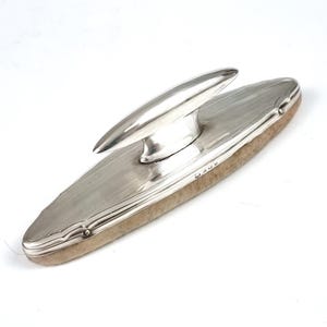 May include: An elongated oval clothes brush featuring a polished silver handle with a subtle engine-turned striped pattern and a central arched grip. The base consists of a dense, light-coloured natural bristle pad with visible hallmarks on the side.