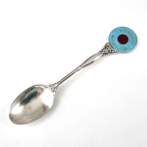 May include: A silver spoon featuring a decorative handle with a circular terminal. The terminal has a light blue enamel border with the text "RINGWOOD L.O.G." and a dark red circular center. The spoon bowl displays clear hallmarks near the stem.