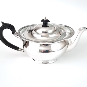 May include: A silver teapot with a black handle and lid detail. The teapot has a curved spout and a decorative rim. The handle is curved and has a decorative design. The lid detail is black and has a rounded shape. The teapot is on a white background.
