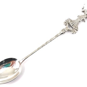 Puede incluir: Una cuchara de plata con un mango adornado con una mujer en un vestido fluido. La cuchara mide aproximadamente 18 cm de largo.