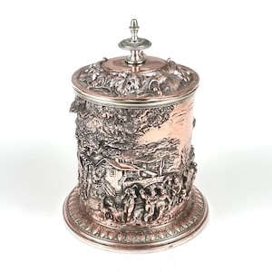 Pode incluir: Um recipiente prateado com tampa, com detalhes em relevo intrincados. A tampa apresenta um design floral e um remate decorativo. O corpo mostra uma cena detalhada de vila com figuras, árvores e uma casa. A base tem uma borda ornamentada e padronizada.