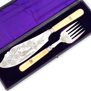 Może przedstawiać: Srebrny zestaw do serwowania z nożem i widelcem w pudełku wyłożonym fioletowym aksamitem. Nóż ma rękojeść z kości i grawerowany wzór ptaka i kwiatów. Widelec ma rękojeść z kości i grawerowany wzór kwiatów.