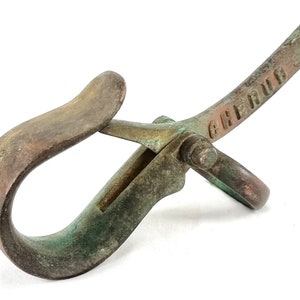 May include: A vintage brass hook with a green patina. The hook has a ring on one end and a lever on the other. The lever is stamped with the word "CHERRY".