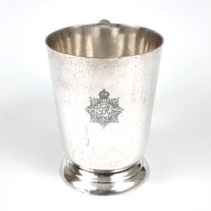 May include: A silver-colored metal cup with a handle and a decorative emblem. The cup has a wide opening and a rounded base. The emblem features a crown and the letters "GR" within a circular design.