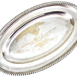 Puede incluir: Una bandeja de servir ovalada de plata con un borde de cuerda. La bandeja tiene una inscripción en letras doradas que dice "The Memorial Trophy of the Tournament of the Champions of the 2nd World's Ladies' Rifle Match, 1926, The Ladies' Rifle Club, London, England".