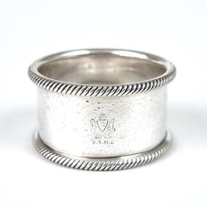 May include: A silver napkin ring with a textured rope design on the top and bottom edges. The ring has a cylindrical shape and features an engraved crest with the letters "O.S.N.C."