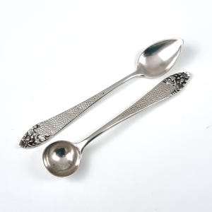 May include: Two silver-toned metal spoons with a polished finish. One spoon features an elongated oval bowl, while the other has a round bowl. Both handles display a textured, hammered surface pattern with detailed floral engravings at the ends.
