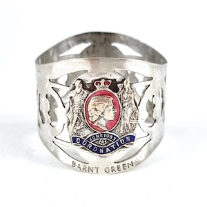 Puede incluir: Anillo de servilleta de plata con un diseño recortado. El anillo presenta un emblema conmemorativo con un retrato de la reina Isabel II y el texto "June 1953 Coronation". El anillo está inscrito con "Barnt Green".