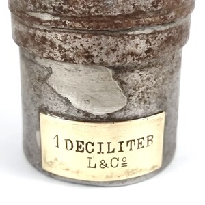 Pu&ograve; includere: Un contenitore in metallo argentato con un'etichetta in ottone che recita "1 DECILITRO L&C&deg;". Il contenitore ha un aspetto usurato e invecchiato.