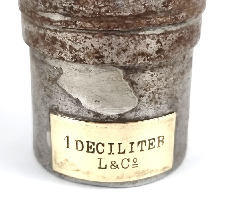 Pu&ograve; includere: Un contenitore in metallo argentato con un'etichetta in ottone che recita "1 DECILITRO L&C&deg;". Il contenitore ha un aspetto usurato e invecchiato.