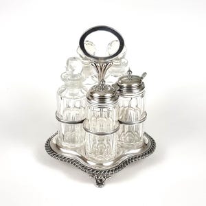 Puede incluir: Un juego de condimentos plateado con frascos y botellas de vidrio transparente. El juego incluye una bandeja, un asa y varios recipientes con tapas y tapones. La bandeja tiene un borde decorativo en forma de cuerda.