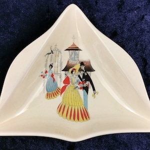 Puede incluir: Un plato triangular de cerámica blanca con un diseño colorido de dos personas con ropa de estilo años 50 caminando bajo un arco floral. El plato tiene un borde elevado y un borde festoneado.