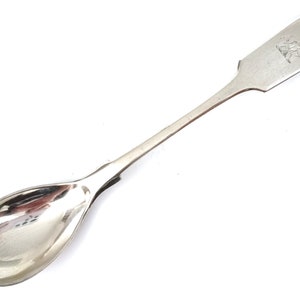 May include: A silver spoon with a rounded bowl and a long, straight handle. The handle is engraved with a design.