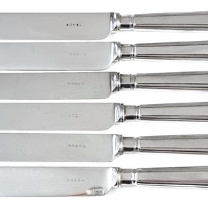 Könnte beinhalten: Satz von sechs silbernen Buttermessern mit verzierten Muschelgriffen. Jedes Messer hat eine glatte, polierte Klinge und einen detaillierten Griff mit einem erhabenen Muschelmotiv.