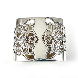 Könnte beinhalten: Ein silberner Serviettenring mit einem verzierten, durchbrochenen Design. Der Metall-Serviettenring weist ein symmetrisches Muster aus wirbelnden Linien und floralen Motiven auf. Das Design wiederholt sich auf beiden Seiten des Rings und schafft einen dekorativen Akzent.