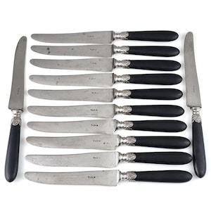 May include: A collection of antique knives featuring silver blades and black handles. The blades are marked "PARIS". Each handle has a decorative silver collar. The knives are displayed on a white background.
