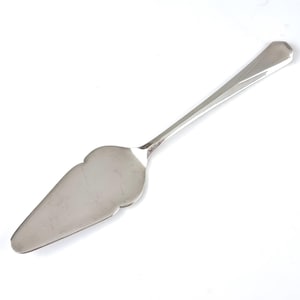 Puede incluir: Un servidor de pastel plateado con un borde decorativo festoneado en la hoja. El mango tiene un diseño geométrico y un extremo plano y angulado. El servidor está hecho de un metal brillante y reflectante.